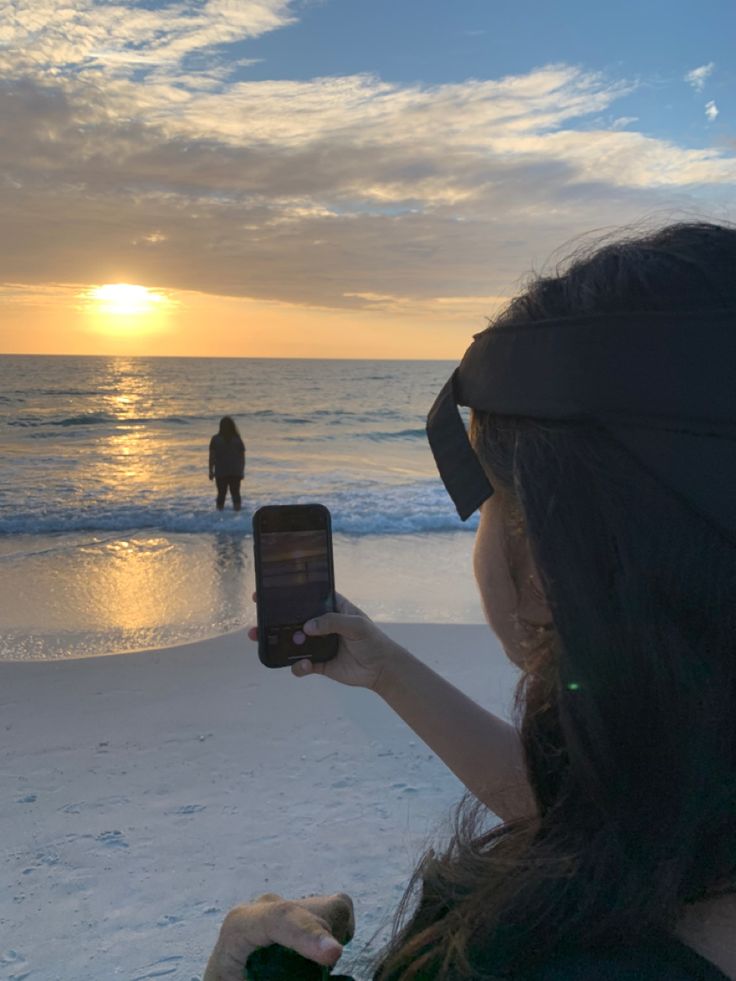 a woman holding up her cell phone to take a photo on the beach at sunset