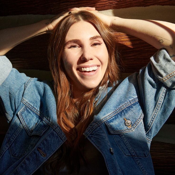 a woman with long hair wearing a jean jacket and smiling at the camera while standing in front of a wooden wall