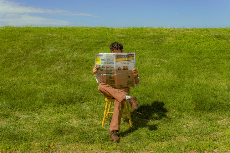 a man sitting on a stool reading a newspaper in the middle of a grassy field