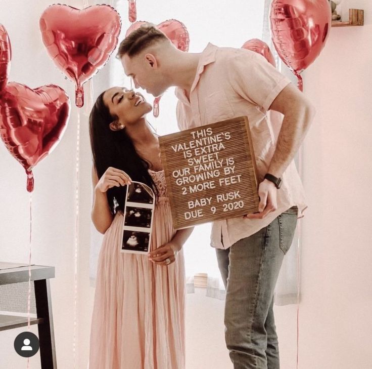 a man and woman standing next to each other in front of heart shaped balloons holding a sign