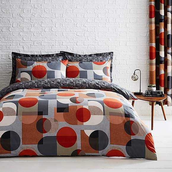 a bed with orange and black comforters in front of a white brick wall next to a window