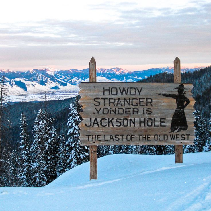 a wooden sign on top of a snow covered slope with trees in the background and mountains in the distance