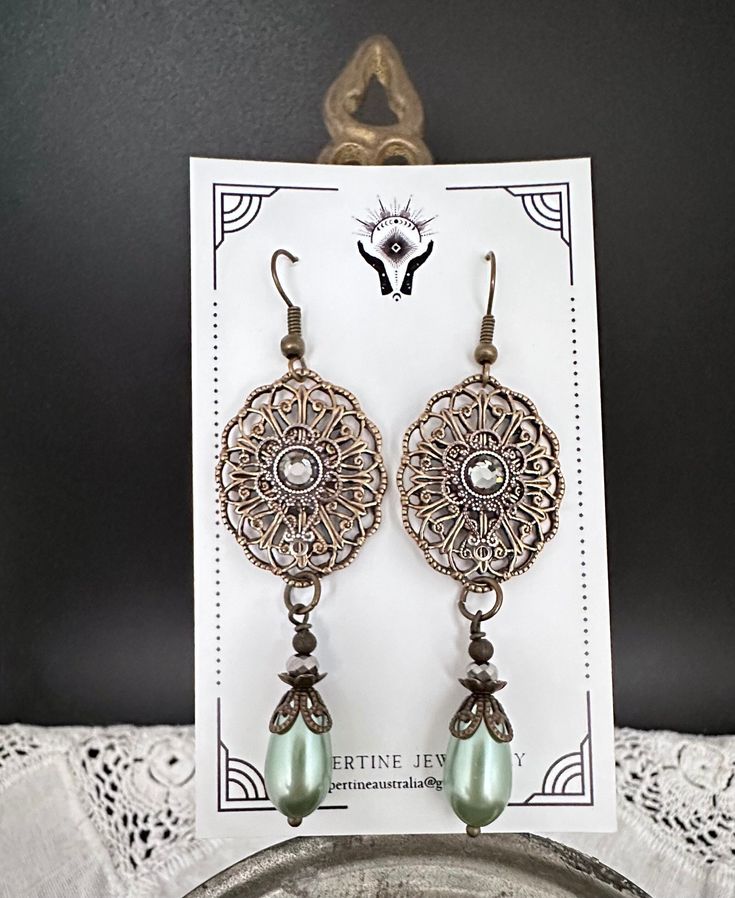 Victorian vintage style filagree earrings with a green pearl drop. The Brass Ox filagree is from USA. I have added a Swarovski flat back to the centre in a silver setting. The drop is a green acrylic pearl which is lightweight and is finished with filagree bead caps, also from USA. The total drop, measured from the top of the earring hook is 70mm or 2 ¾ inches. The earring hooks are antique bronze, and are nickel free. ♥︎ BRASS OX COMPONENTS Is the colour of old antiqued brass.  The plating is a professional process, commercially applied by the finest costume jewellery brass finishers in the business, according to strict industry standards for jewellery components. These Filagree's and Stampings will last for years with simple care. Many of the pieces are still made from the original dies Green Pearl Earrings, Red Stone Earrings, Coin Pearl Earrings, Large Drop Earrings, Grey Pearl Earrings, Patina Earrings, Moon And Star Earrings, Wedding Bridesmaid Jewelry, Earring Dangle