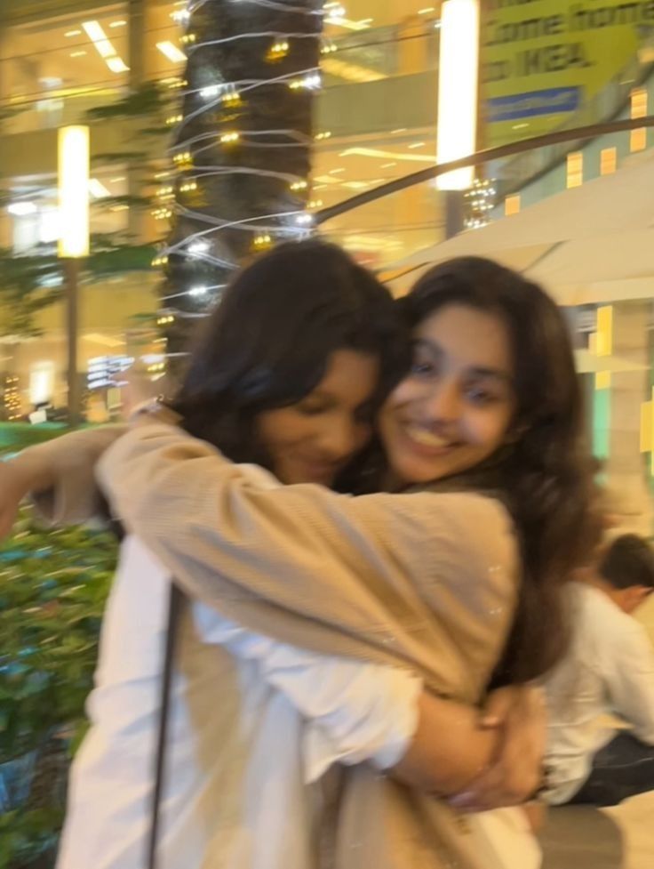 two young women hugging each other in front of a building with lights on the ceiling