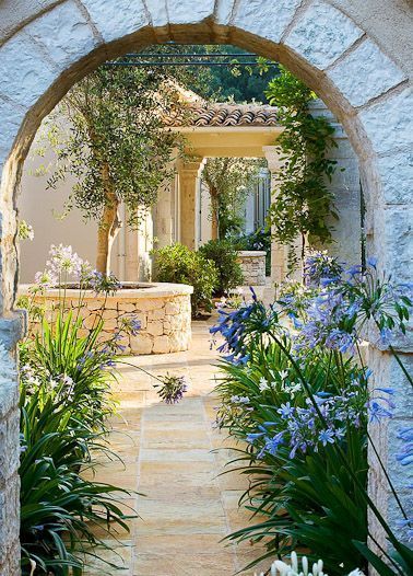 an archway leading to a house with blue flowers and greenery in the foreground