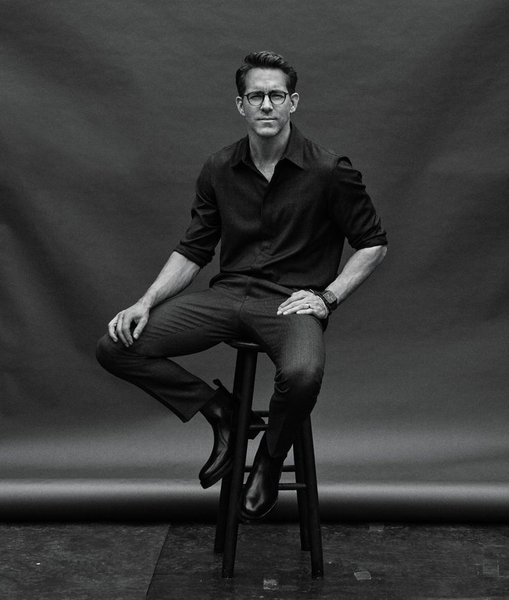 a man sitting on top of a wooden stool in front of a gray background wearing glasses