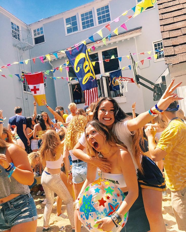 two young women are hugging and having fun at a party with other people in the background