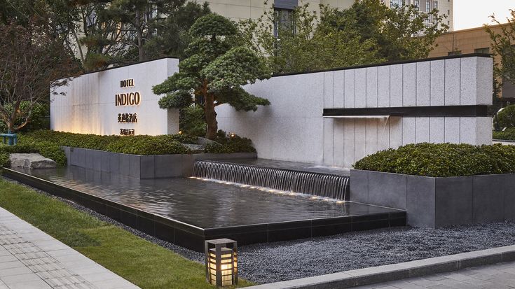 a water feature in front of a building with trees and bushes around it on the side walk