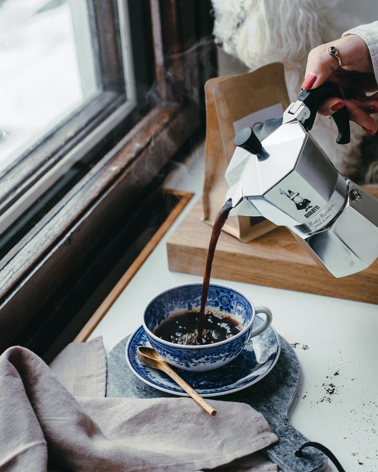 a person pours coffee into a blue cup on a table next to a window