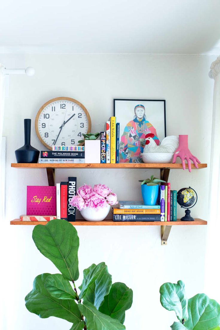 two wooden shelves with books, plants and a clock on the wall above them in a living room