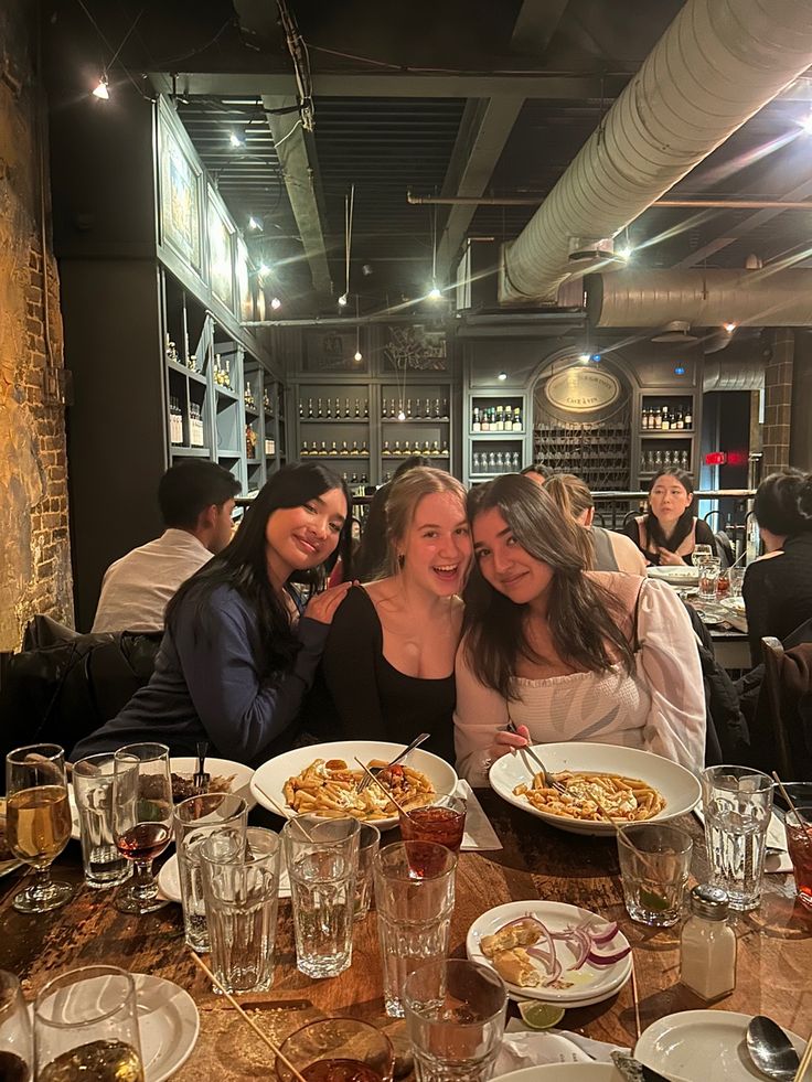 three women sitting at a table with plates of food in front of them and wine glasses on the table