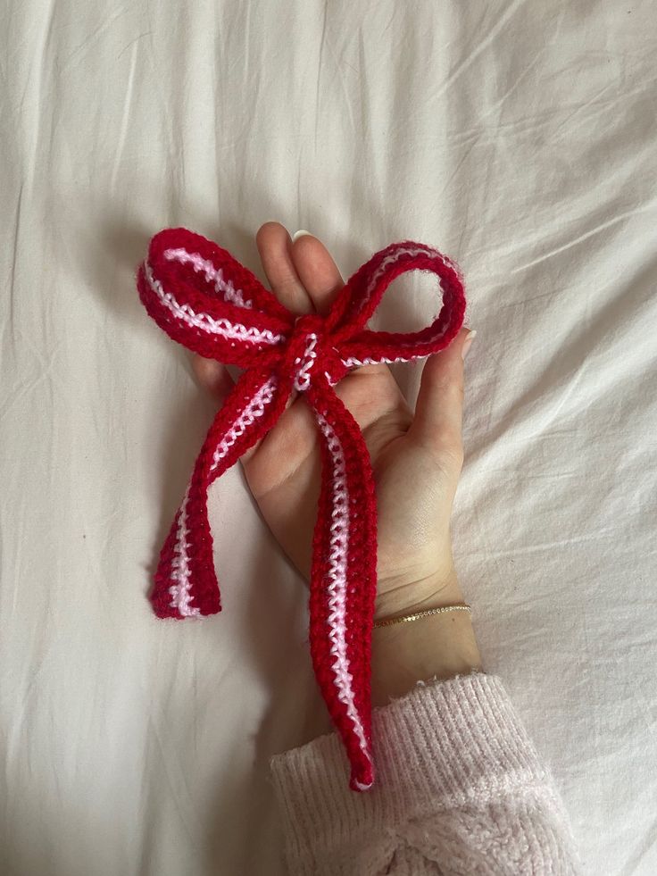 a hand holding a red and white crocheted bow on top of a bed