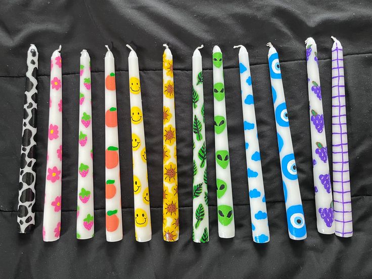 six different colored candles are lined up on a black surface, with faces drawn on them