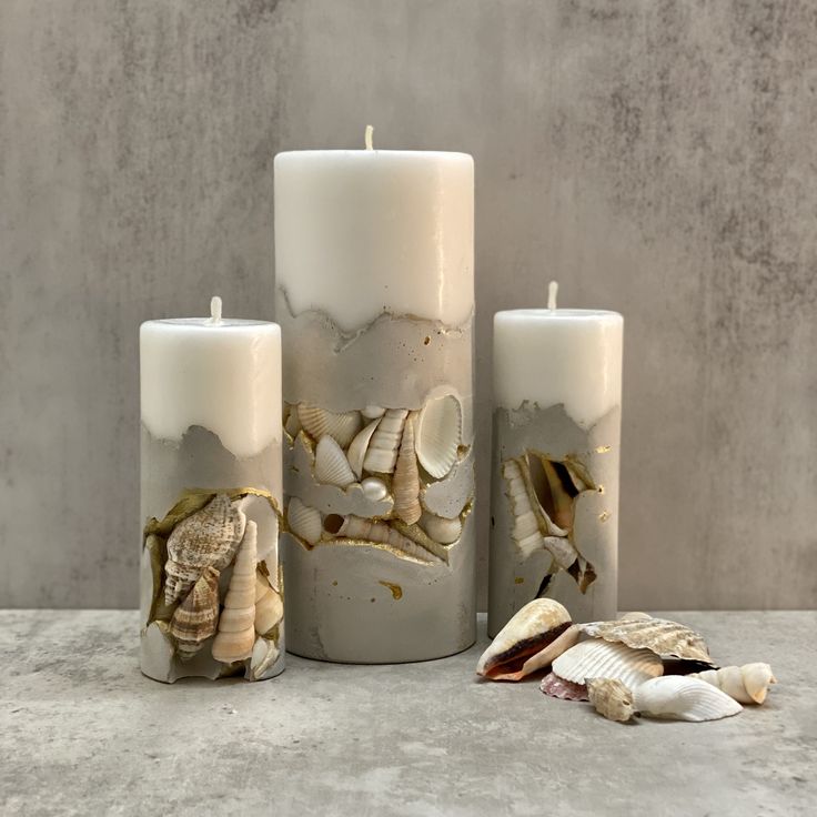 three candles with seashells on them sitting next to each other in front of a concrete wall