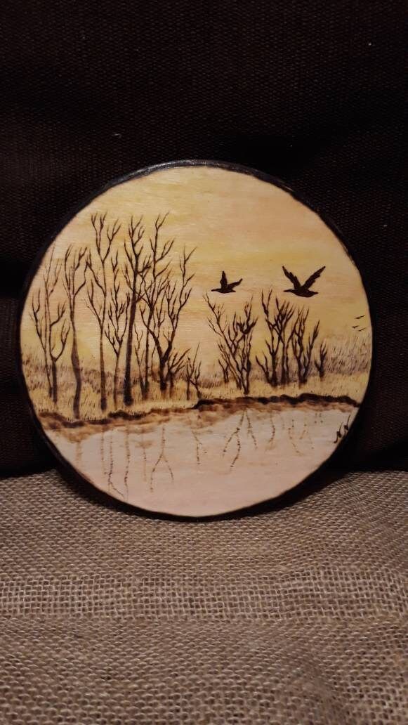 a wooden plaque with trees and birds flying over the water in front of a brown background