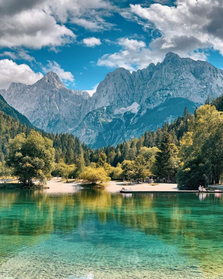 the mountains are reflected in the clear blue water on the lake's surface, while trees and bushes stand along the shore