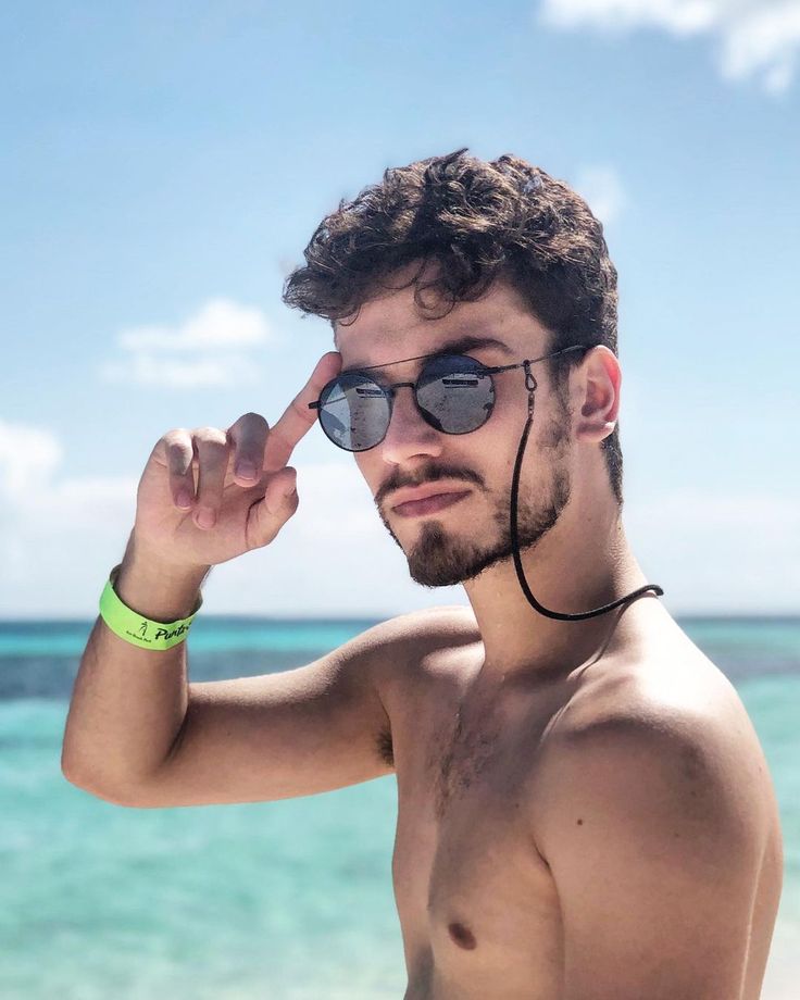 a shirtless man with sunglasses on the beach looking at the camera while holding his hand up to his eye