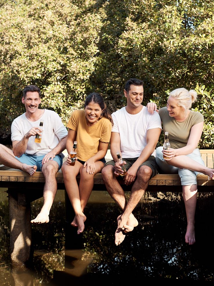 four people are sitting on a wooden bench by the water and holding beers in their hands