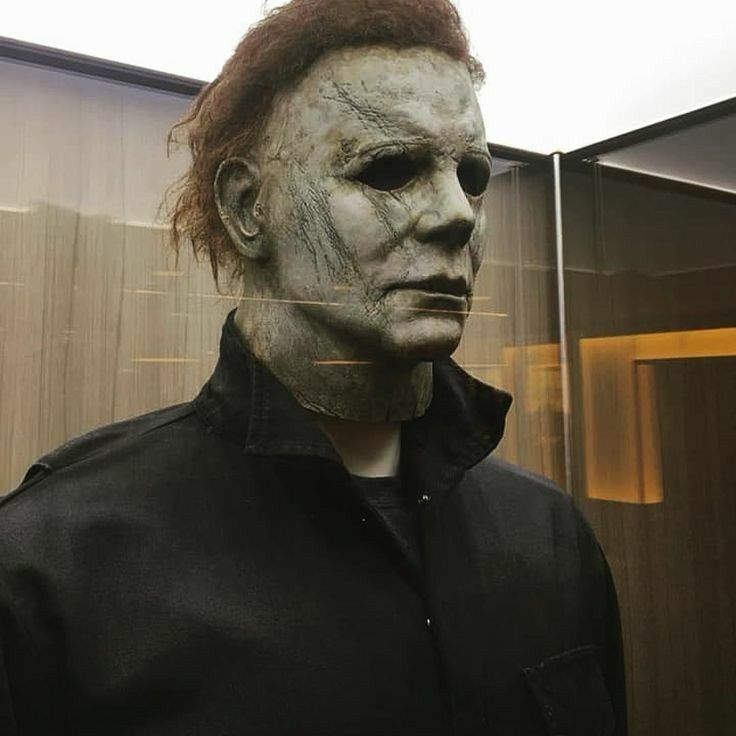 a man in a black shirt wearing a creepy mask and looking at the camera while standing next to a mirror