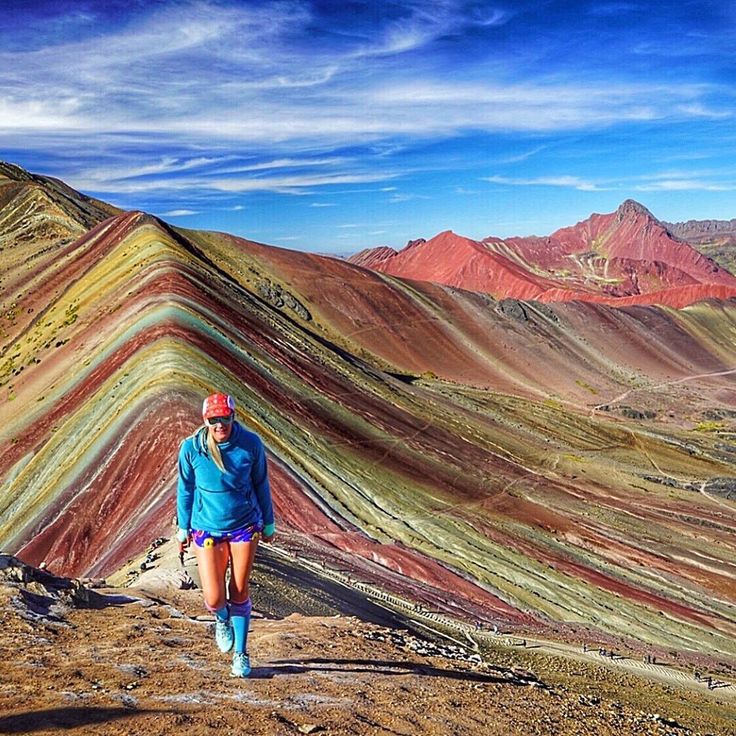 a woman in blue jacket standing on top of a hill with colorful mountains behind her