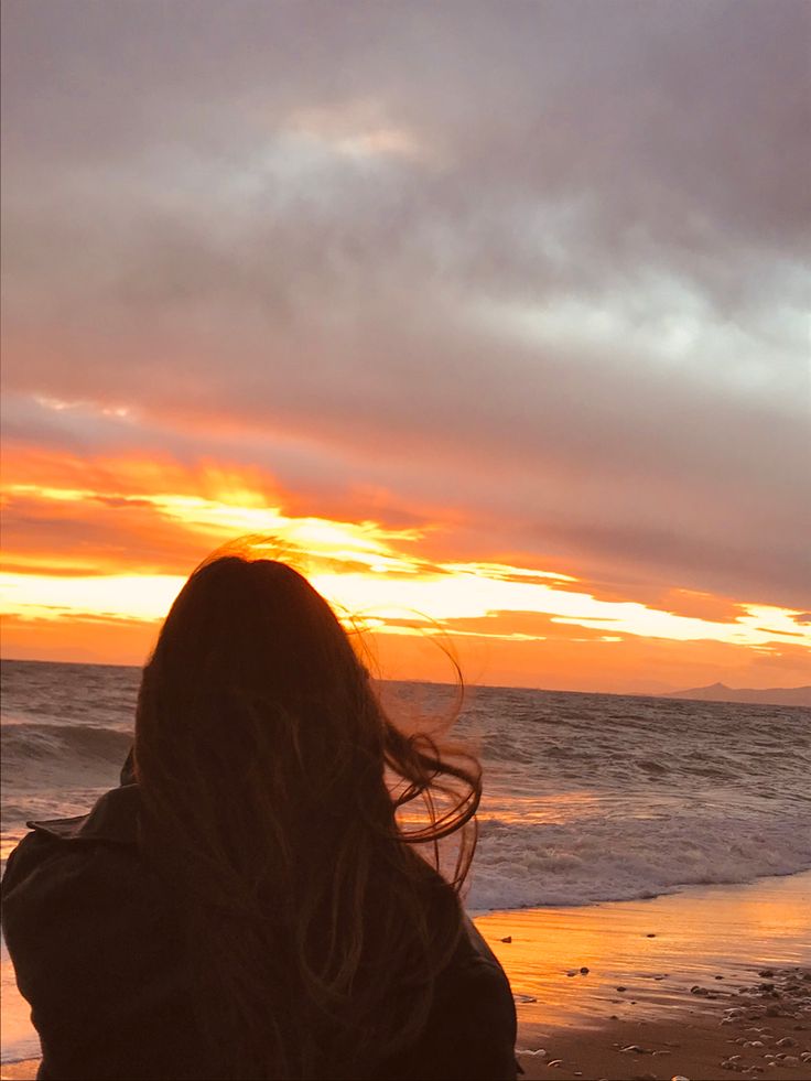 a woman standing on top of a sandy beach next to the ocean under a cloudy sky