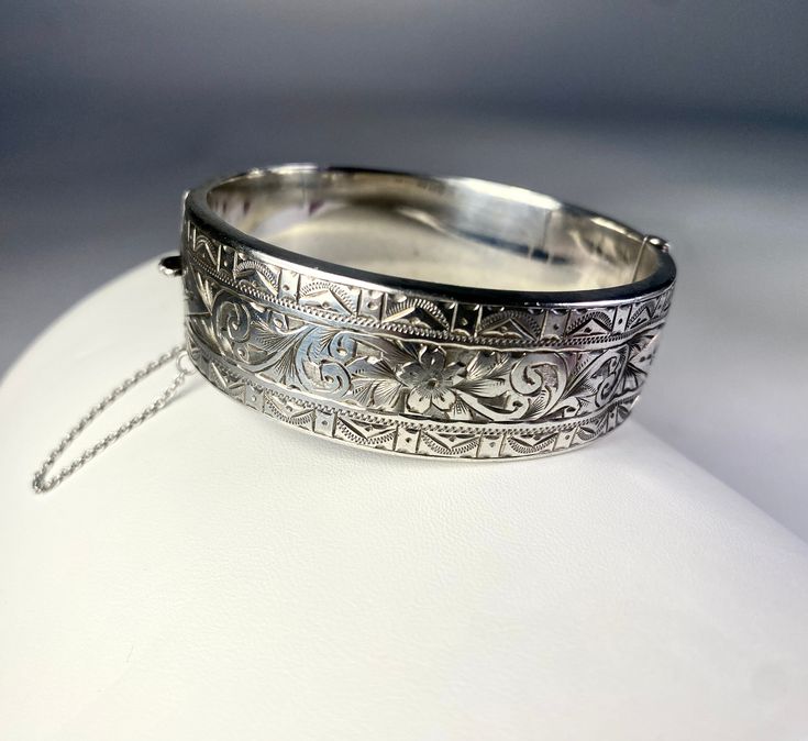 "Sterling Silver Bangle Bracelet with fine engraving. The design is boarded top and bottom of engraving in a triangular geometric  pattern. The interior panel is a floral motif. There is a dent in the panel but it doesn't take away from this beautiful piece with  history. The box closure works well and there is a safety chain.  Size: 3/4\" Width, Interior diameter 2 3/8\"  Thank you visiting HATTIE MAUDE VINTAGE  and please reach out with any questions, we are here to help!" Luxury Vintage Engraved Bangle, Luxury Ornate Sterling Silver Bangle, Luxury Antique Sterling Silver Bangle Bracelet, Formal Engraved White Gold Bangle, Antique Round Cuff Bracelet For Wedding, Antique Cuff Bracelet With Intricate Design For Formal Occasions, Victorian Etched Cuff Bracelet, Victorian Silver Engraved Bangle, Victorian Etched Cuff Bracelet For Wedding