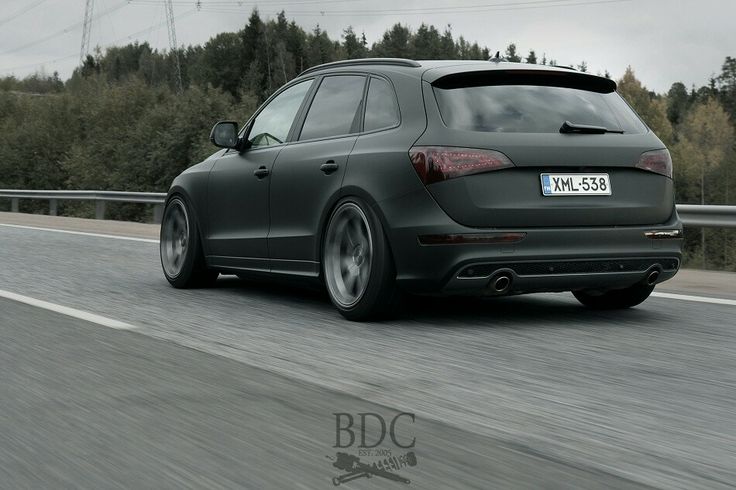 the rear end of a green car driving down a road with trees in the background