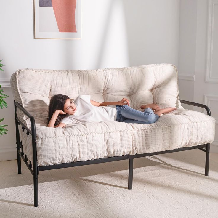 a woman laying on top of a white futon couch in a living room next to a potted plant