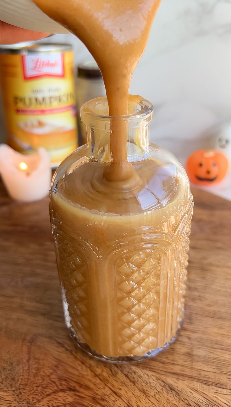 a person pouring caramel into a glass jar on a wooden table with pumpkins in the background