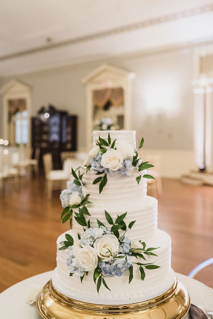 a white wedding cake with blue flowers and greenery on top sits on a gold platter