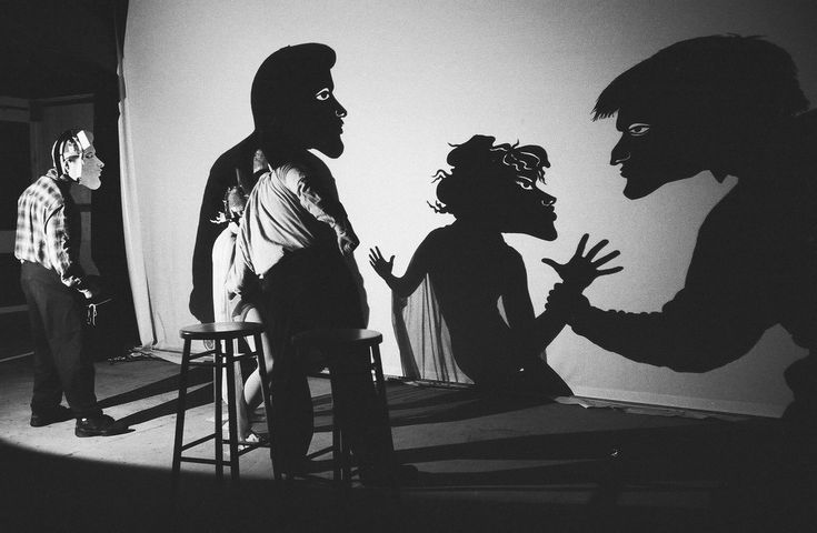 three people standing in front of a white wall and one person sitting on a stool