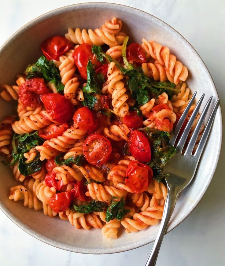 pasta with tomatoes and spinach in a white bowl next to a fork on a marble surface