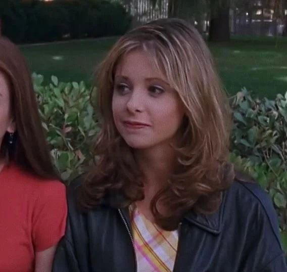 two women standing next to each other in front of some bushes and trees with one woman looking at the camera