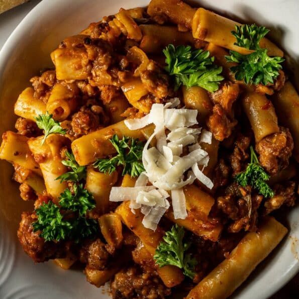 a white plate topped with pasta and meat covered in sauce, parmesan cheese and parsley