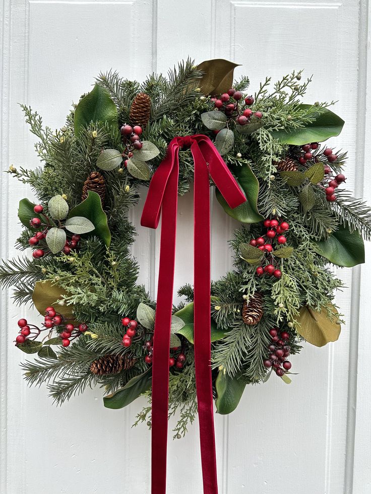 a christmas wreath with red ribbon hanging on the front door, decorated with greenery and pine cones