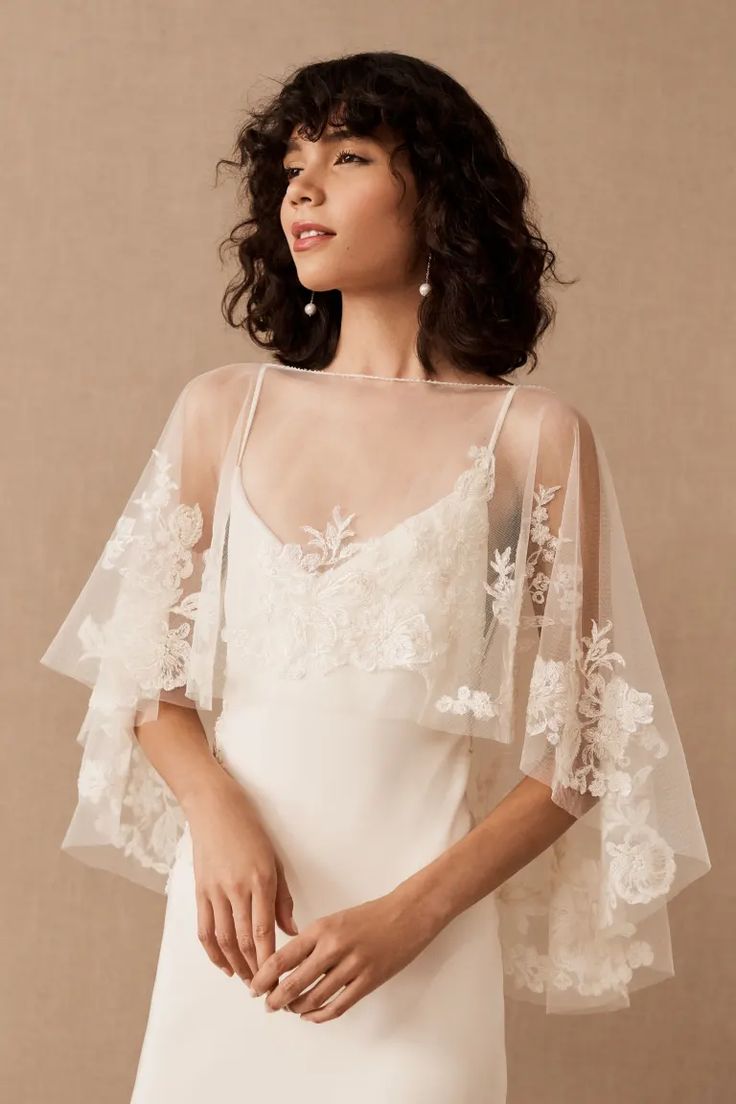 a woman wearing a white dress with sheer sleeves and flowers on the shoulder, standing in front of a beige background