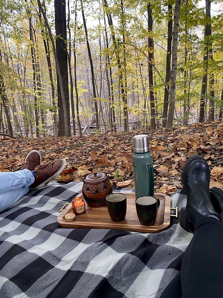 a person laying on a blanket in the woods with their feet propped up next to coffee cups