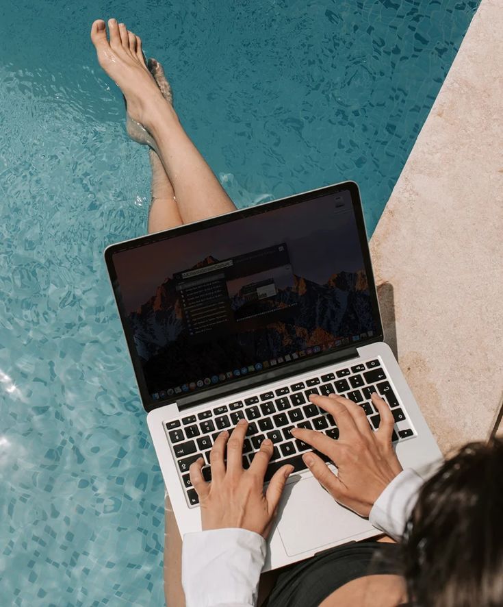 a person sitting in front of a swimming pool using a laptop with their feet on the edge