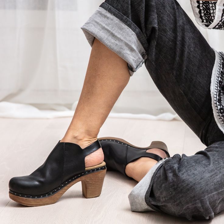 Introducing the Tyra slingback clog. This high-heeled clog in buttery soft black leather will elevate any look. Handcrafted in Portugal with meticulous attention to detail, the Tyra clog features a rounded toe, visible rivets, and an adjustable ankle strap with a gold buckle for a custom fit. The wood-like heel marries style with comfort, ensuring you can step out in style, no matter the occasion. Heel height 7,5 cm Normal fit Black Clogs With Sculpted High Heel, Black Closed Toe Clogs With Sculpted Heel, Chic Clogs With Heel Strap And Open Heel, Chic Block Heel Clogs With Heel Strap, Black Clogs With Wooden Heel For Work, Spring Slingback Clogs With Heel Strap, Black Workwear Clogs With Wooden Heel, Chic Clogs With Wooden Block Heel, High Heel Clogs With Leather Sole For Work