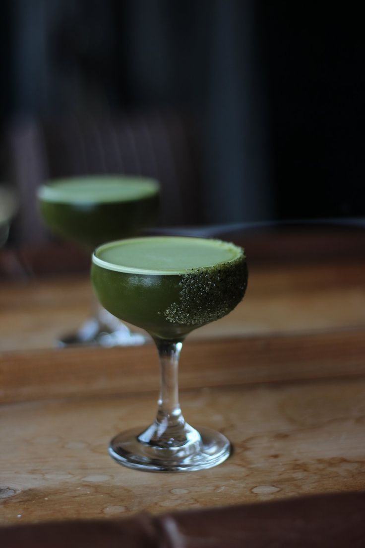 two glasses filled with green liquid sitting on top of a wooden table next to each other