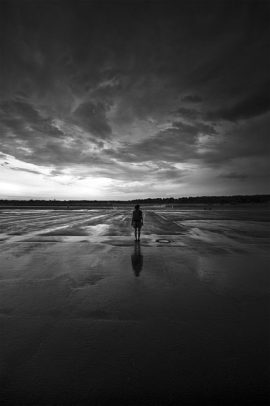 black and white photograph of person walking on wet beach at dusk with dark clouds in the background