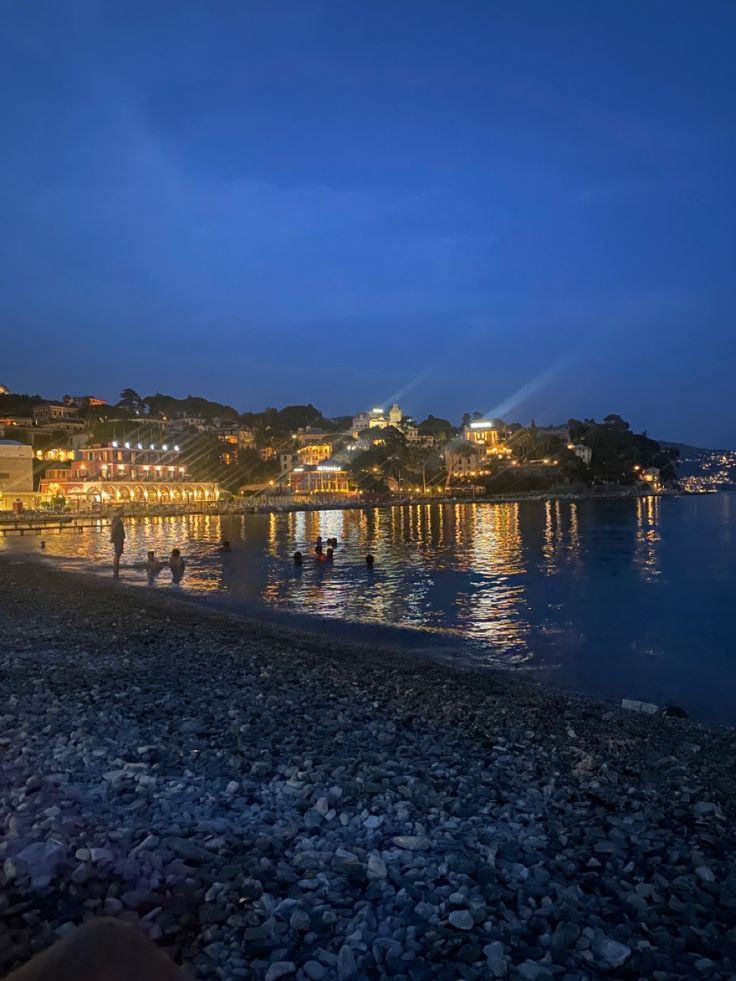 people are sitting on the beach at night with lights shining in the water behind them