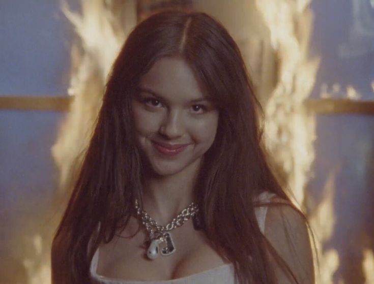 a woman with long brown hair wearing a white top and necklace smiling at the camera