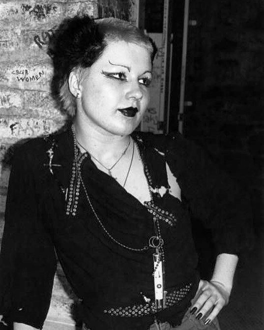 a woman with black makeup and piercings standing next to a brick wall