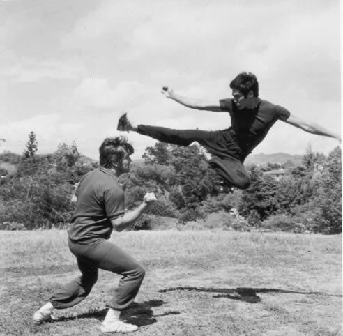 two men are playing baseball in an open field with trees and bushes behind them, one man is jumping to catch the ball