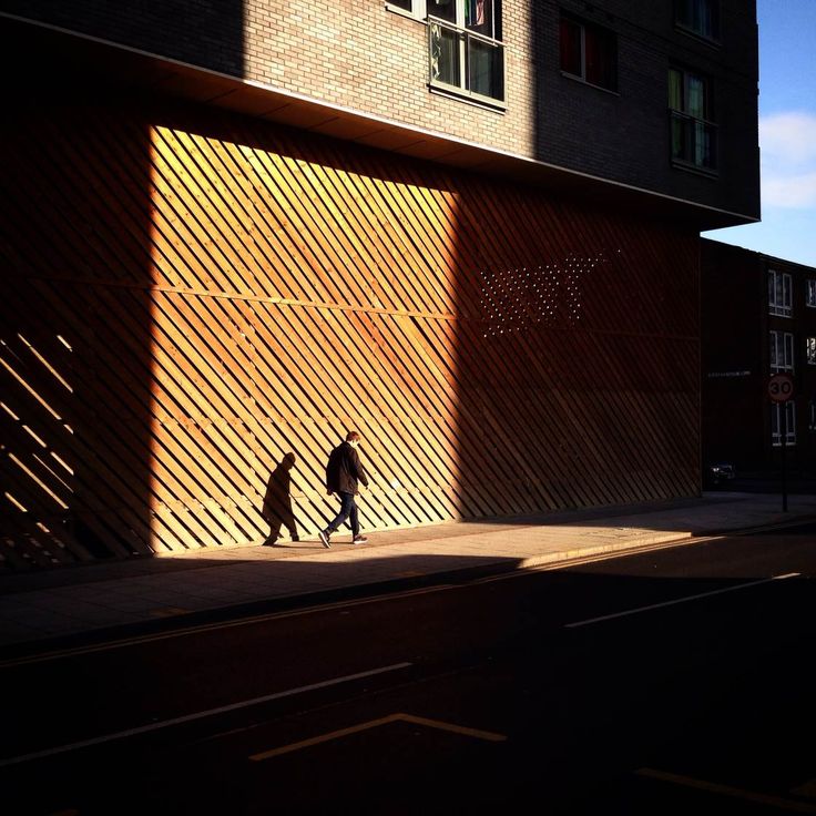 two people are walking down the street in front of a building with shadows on it