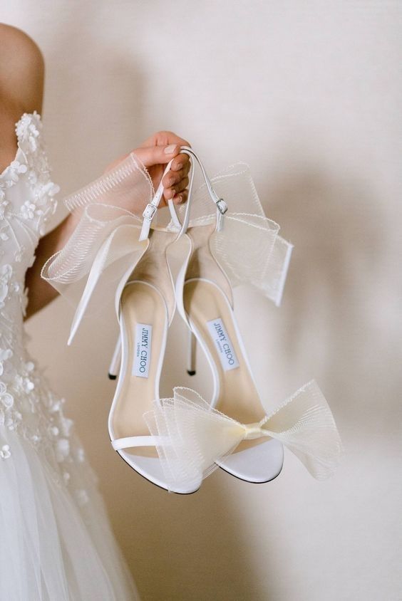 a woman in a wedding dress holding up her white high heeled shoes with bows