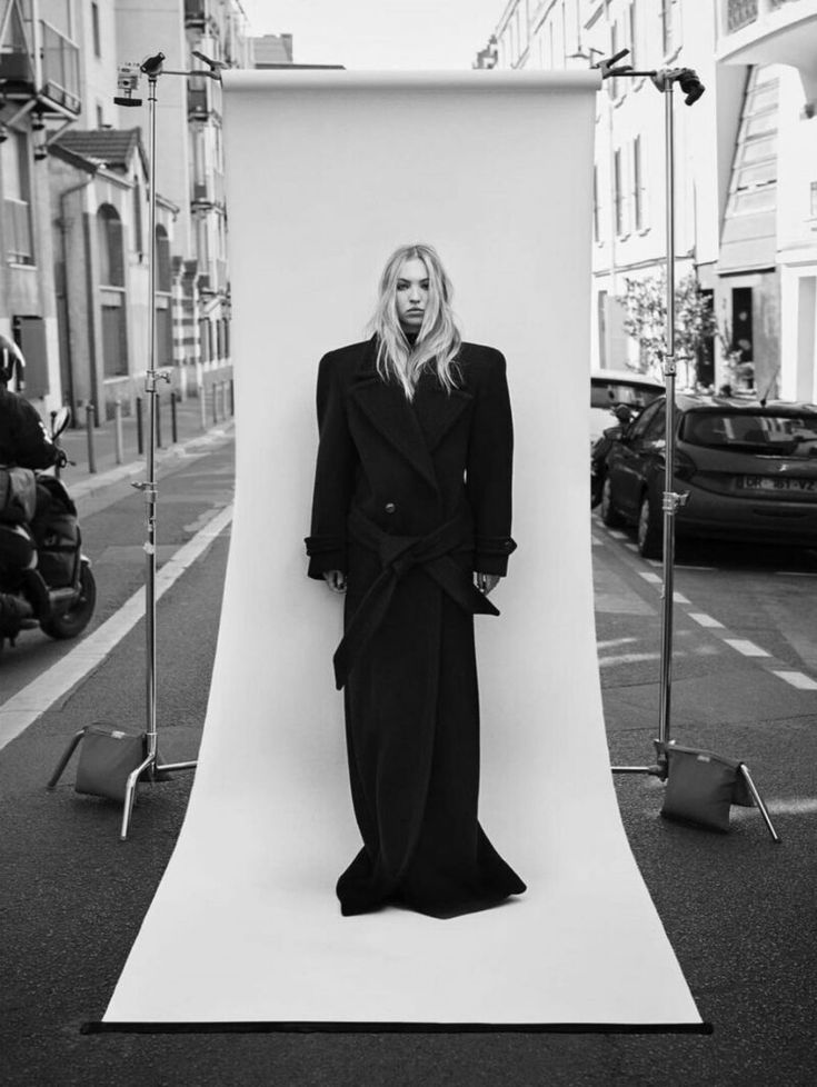 a woman standing in front of a white backdrop with her hands on her hips, wearing a long black coat and pants