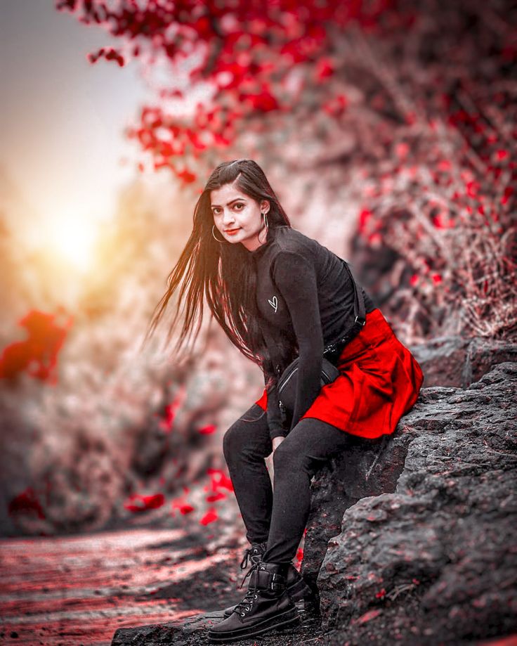 a woman with long hair is sitting on a rock and posing for the camera in front of red leaves