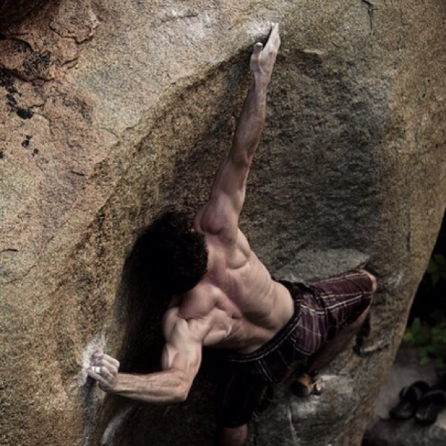 a shirtless man climbing up the side of a large rock with his hands in the air